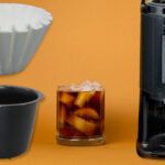 The image shows coffee filters, a black brewe basket, a glass of cold brew with icecubes (symbolizing the cold brew function) and a 2.5l thermos. All these articles are included in a  starting kit basic