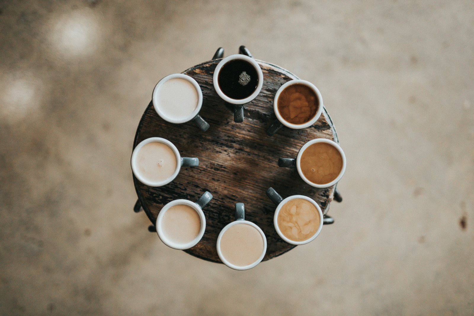 This image captures a bird’s eye view of a rustic wooden table adorned with eight distinct mugs of coffee. Each mug, filled with a unique brew, is arranged in a perfect circle, showcasing the diversity and artistry in coffee preparation. The image serves as a visual delight for coffee enthusiasts, highlighting the variety and richness of different coffee brews.